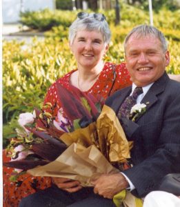 Ray in a suit sits outdoors with bouquet and Linda Leonard