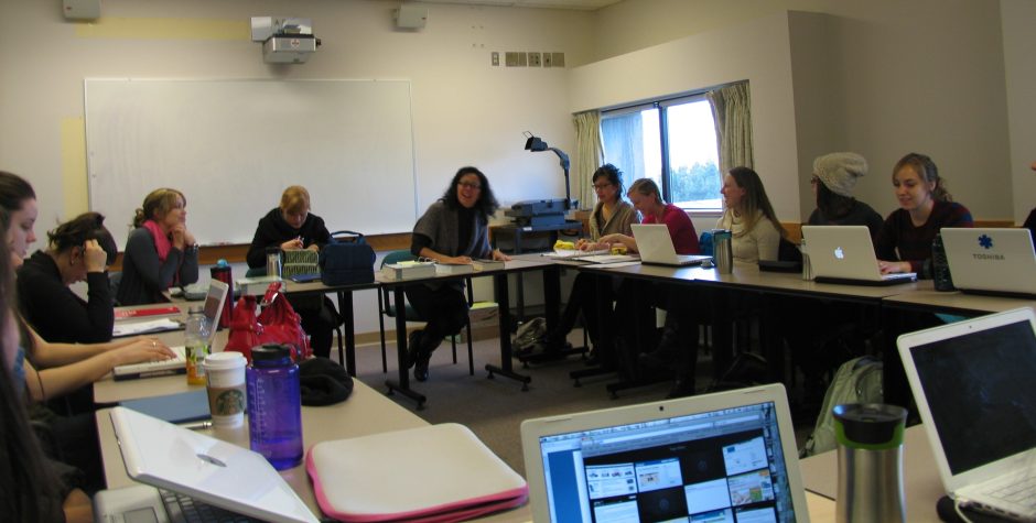 Elsie sits at a table with a group of students who are seated in a classroom with the desks arranged in a circle, each with a laptop.